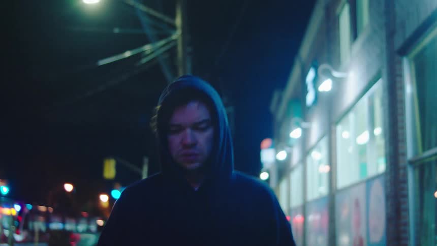 The man in the blue jacket is walking down the sidewalk and continues to look behind him and has many nervous signs like rubbing his face and headhe is acting as if someone might be following him