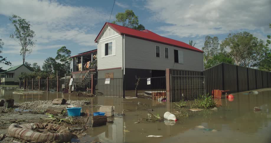 A raised house with a red roof sits surrounded by muddy floodwaters and scattered debris