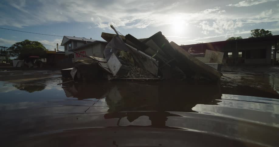 A large pile of flood damaged items sits in muddy water reflecting the bright sky with the sun shining behind it and damaged buildings in the background