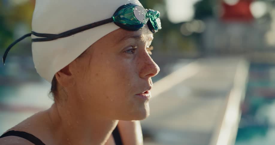 A close view shows a woman wearing a white swim cap and mirrored green goggles sitting next to a pool breathing heavily in the bright afternoon sunlight