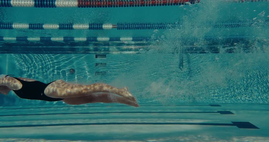 A swimmer wearing a black swimsuit executes the freestyle stroke beneath the waters surface propelling herself through a clear sunlit competition pool with visible lane lines