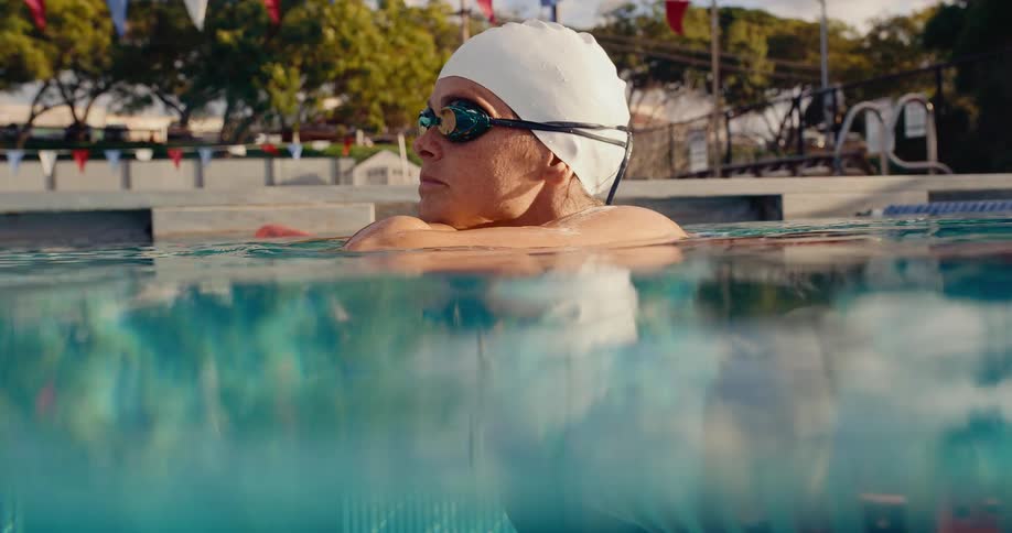 Swimmer with white cap resting on lane divider in pool with flags and trees in background