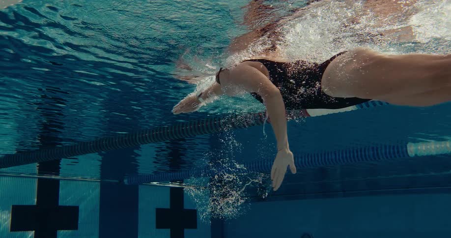 Young adult female wearing a black swimsuit with a swim cap and goggles swimming on a pool during day time