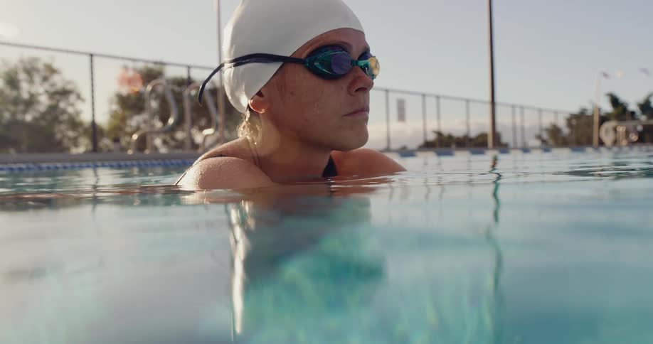 Swimmer with white cap and blue goggles remains partially submerged in pool beside metal ladder