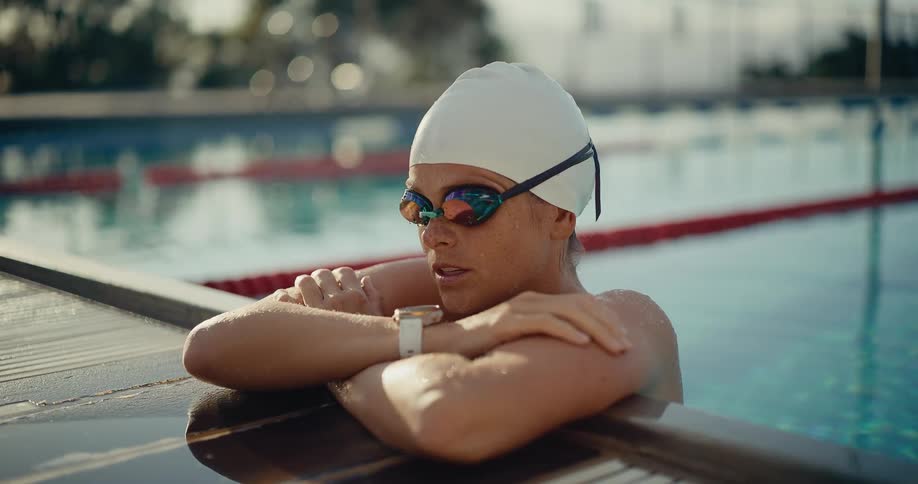 A young woman rests in the swimming pool with her hands on the pavement wearing a swimming hat and goggles as she looks at her wristwatch on a sunny day