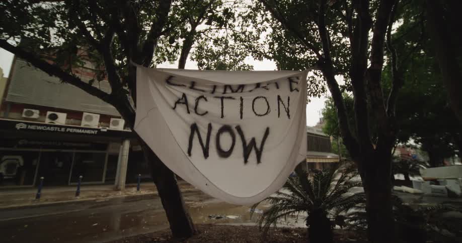 A white cloth banner with the words climate action now spraypainted in black is tied between two trees waving gently in the wind over a wet quiet urban streetscape on a rainy day