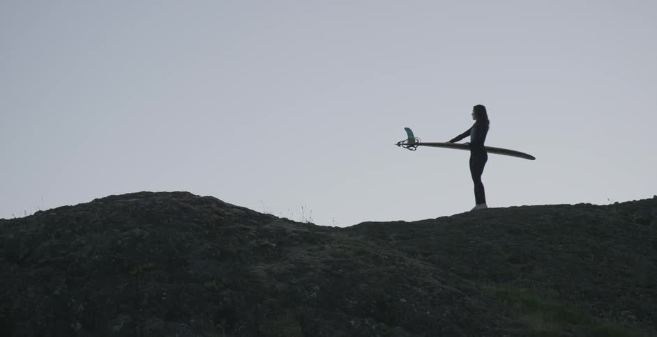A surfer stands with a surfboard on a hill