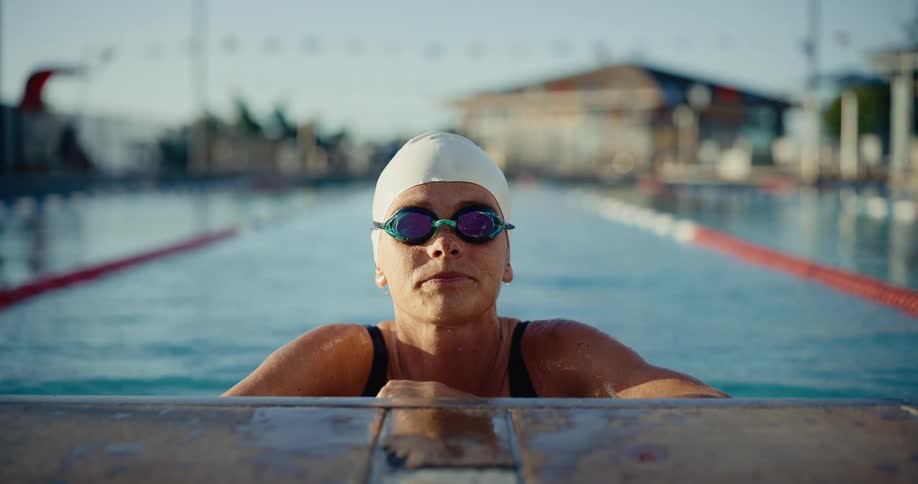 Young adult woman wearing a bathing suit and swim cap with goggles resting at the edge of a swimming pool outdoors during day time