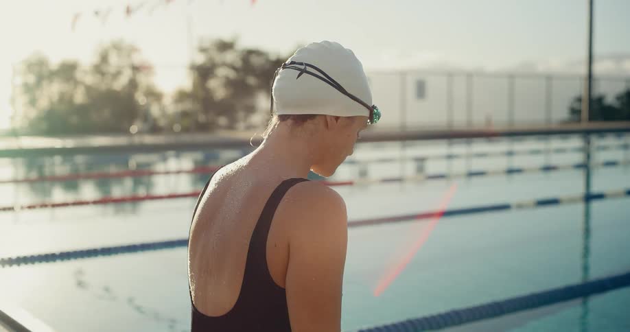 Person wearing white cap and black swimsuit facing outdoor pool with lanes and trees in background