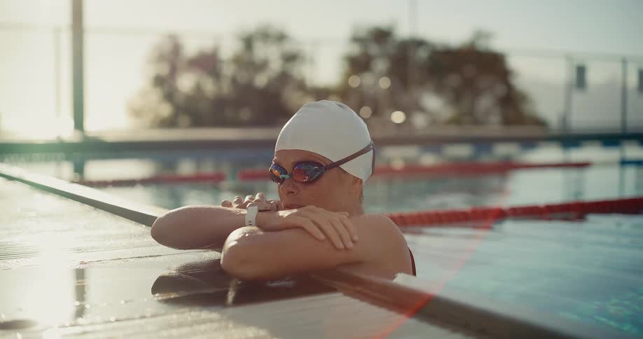 A female swimmer wearing a white cap and mirrored goggles resting her forearms on the edge of an outdoor pool looking intently into the suns bright lens flare