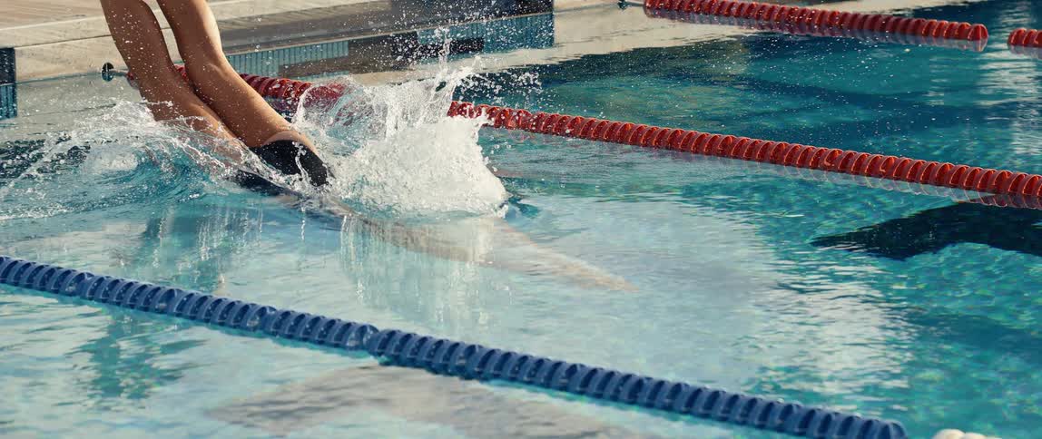 Young female adult wearing a black swimming suit and a swim cap diving into a pool and swimming outdoors during day time