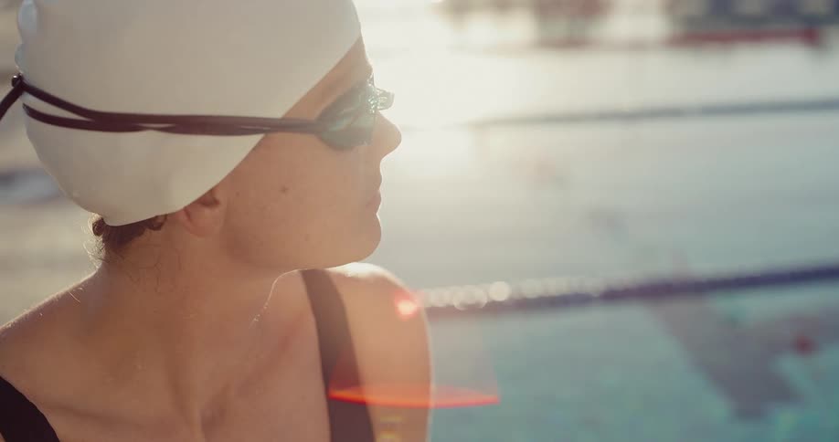 A closeup shot focuses on a swimmer wearing a white cap and mirrored goggles sitting by the edge of an outdoor pool the bright sunlight reflects off the water creating a lens flare and highlighting the swimmers profile as she looks out over the water