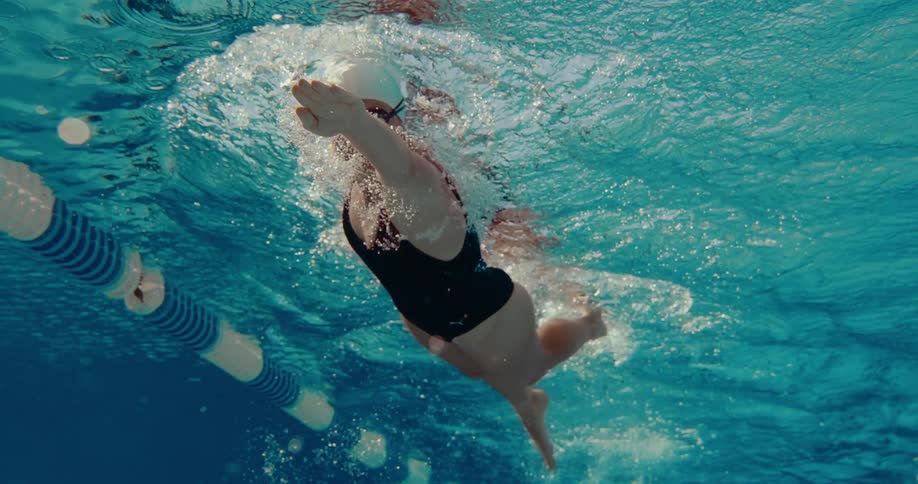 An underwater shot captures a female swimmer moving in a black swimsuit and white cap performing the freestyle stroke across a bluetiled pool lane