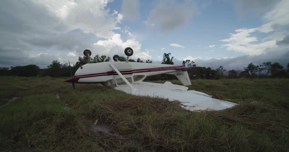 This video shows a small fixedwing aircraft lying upside down in a grassy field suggesting a crash or emergency landingthe white plane with maroon stripes has its landing gear and registration number 8bhbh visible against a partially clou