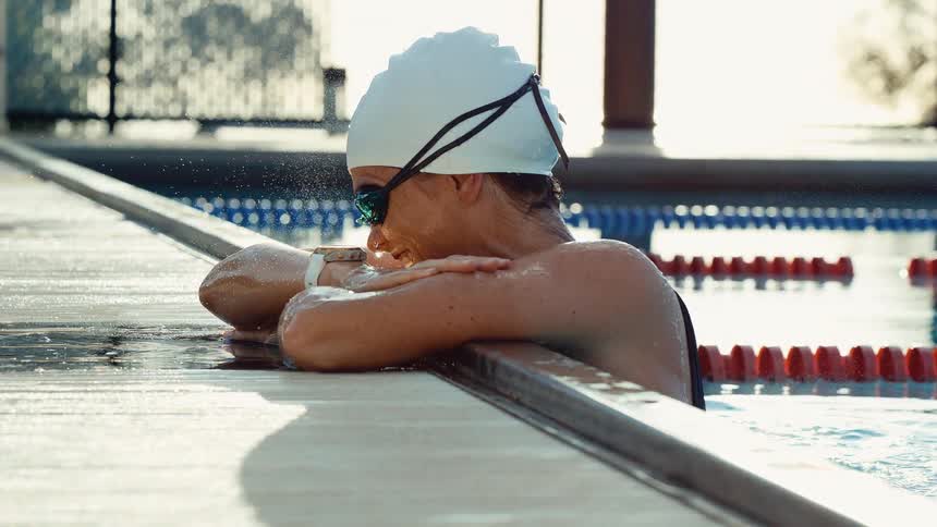 A swimmer wearing a white cap and goggles rests their forearms on the edge of an outdoor pool looking down at the water with a satisfied expression the bright sunlight reflects off the water and highlights the water droplets on their face giving the