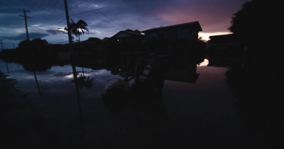 Boat with person floats along flooded street between houses reflected in water at dusk