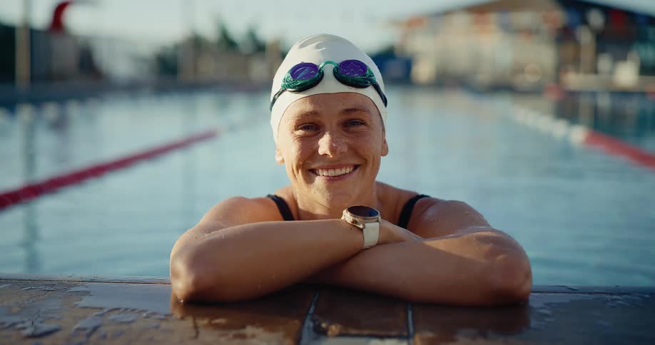 A female swimmer with a swim cap and goggles smiles brightly resting her arms on the edge of an outdoor swimming pool on a clear sunny day with a smartwatch visible on her wrist