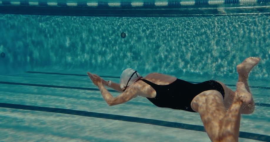 An underwater view shows a swimmer in a black swimsuit and white cap gliding through a lane in a pool the water is clear and the sunlight creates a shimmering pattern on the pool floor as the swimmer performs a pushoff and glide