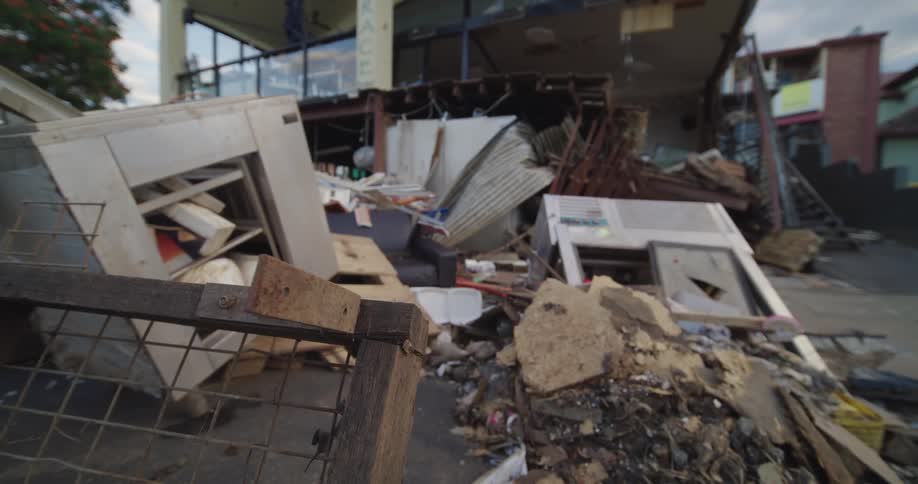 Piles of flood debris and rubble including broken cabinets and metal sheets sit on a muddy street in front of a severely damaged commercial building