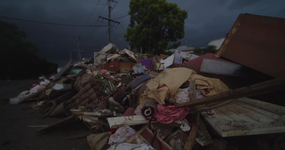 A large pile of debris including mud covered furniture and building materials sits on a street under a dark cloudy sky after a flood or storm