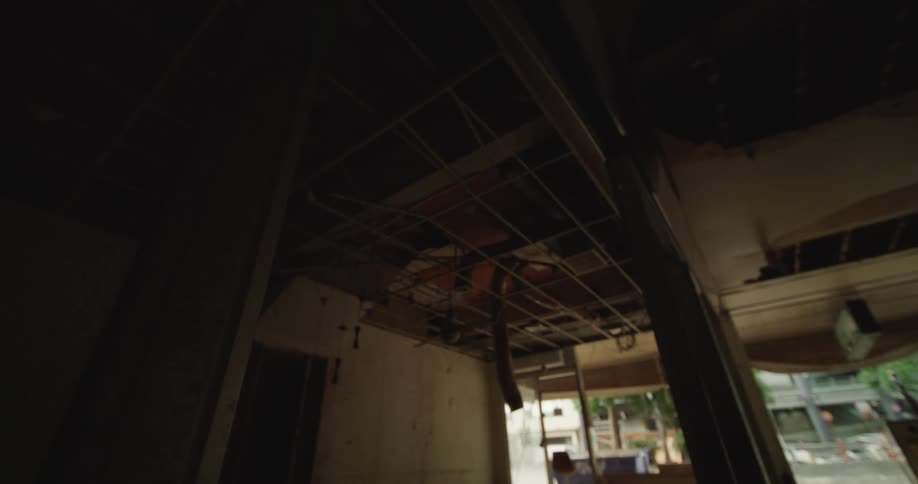A low angle interior view of a dilapidated dark room showing the exposed metal grid of a dropped ceiling and damaged walls with an outdoor view visible through an opening