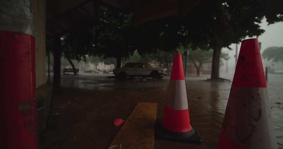 Two orange and white traffic cones stand on a wet covered sidewalk looking out at heavy rain falling on a street with dark trees and a parked vintage car