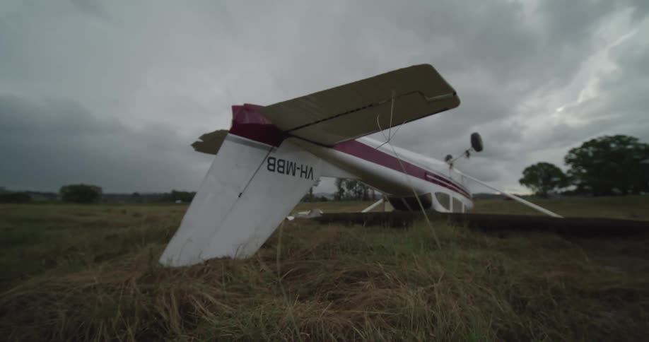 Small airplane lying upside down in a field under a cloudy sky as light traffic passes in the distance