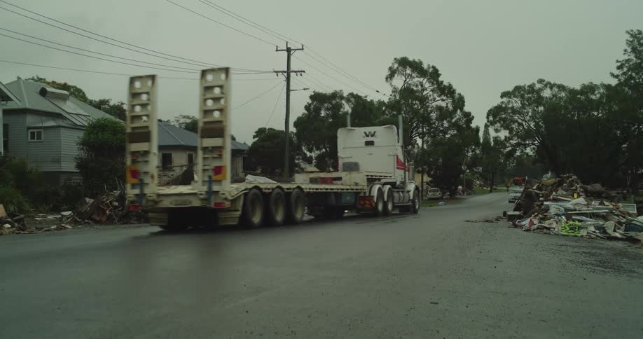 A street in a residential area is lined with massive piles of flooddamaged household debris on both sides on a rainy overcast day also a large semitrailer truck and a sedan pass through the cleared center of the road