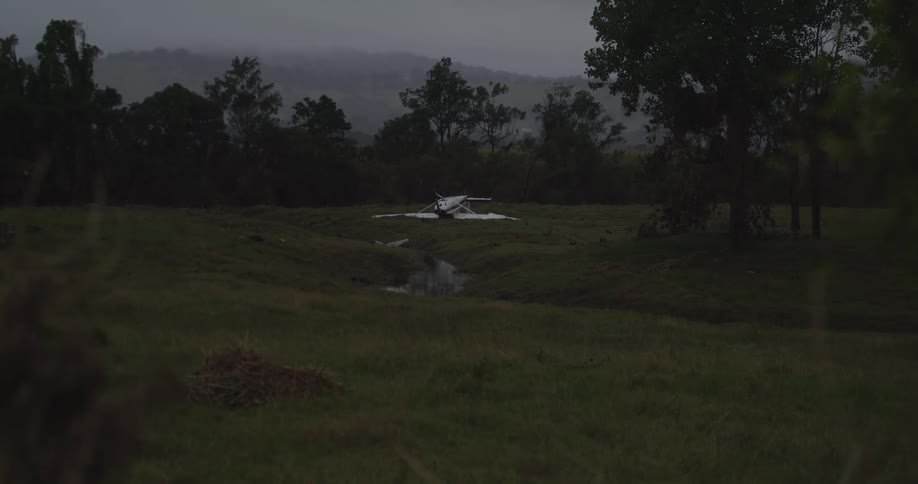 Broken white plane lies crashed in a grassy field surrounded by trees and hills under a cloudy sky
