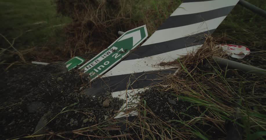 A destroyed road sign pointing left lays broken on the ground next to a striped traffic marker surrounded by wet dirt and tall grass