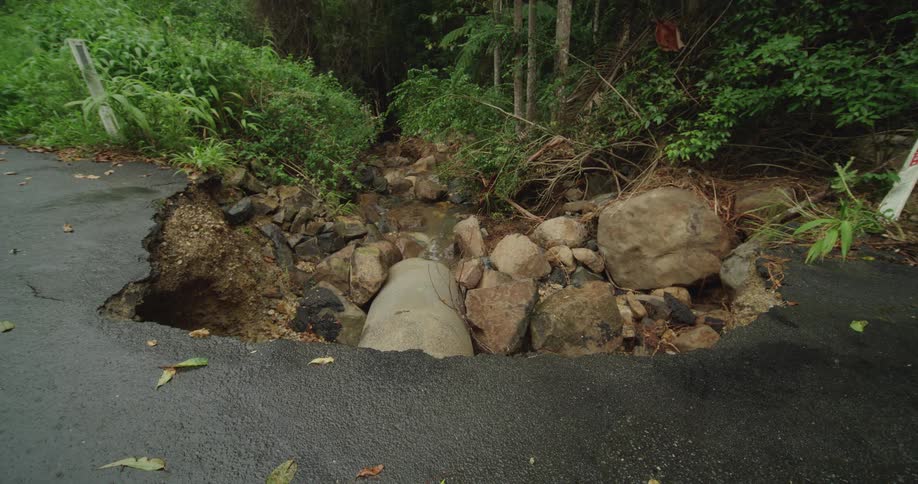 A pothole on the side of the road is covered in rubble and rocks with grasses along the roadside
