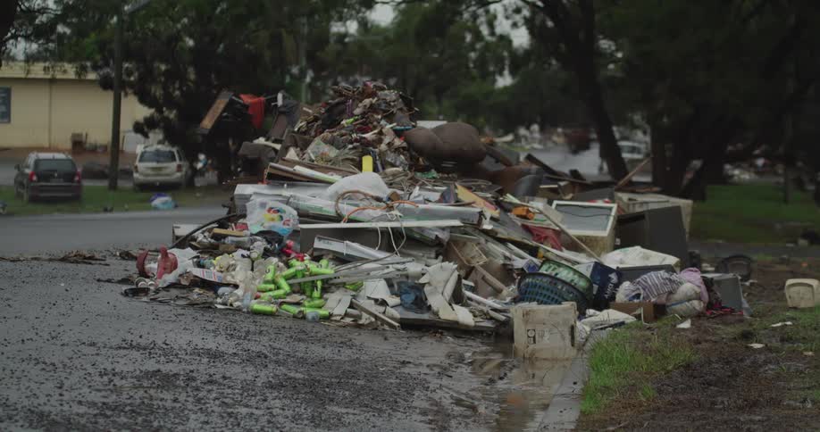 An enormous chaotic mound of flood debris household trash and discarded furniture overflowing onto the wet asphalt and curb of a suburban street with cars passing slowly in the background under an overcast sky