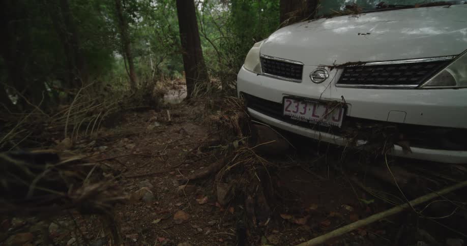 This short video shows the front end of a white nissan sedan stuck in a heavily debrisfilled area likely after a flood or mudslidethe car is wedged against a tree surrounded by mud sticks and roots with its queensland australia license plate