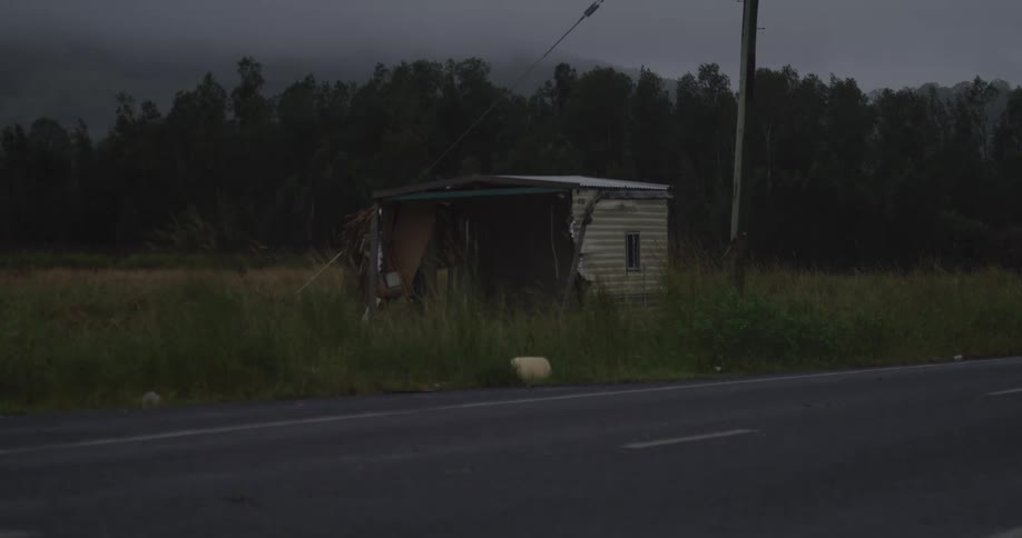 This video features a very dark and dilapidated corrugated metal shack or shed next to a paved road possibly an abandoned trailera few vehicles drive past in the foreground while a dense line of dark trees and a low mountain or hillside are visible u