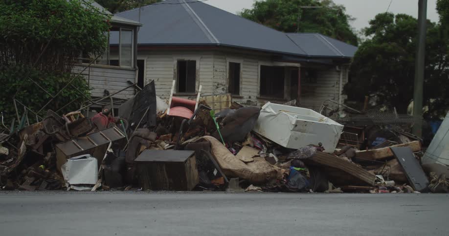 A pile of discarded furniture sits outside a building by the roadside at midday with cars passing by and a help sign placed among the trash
