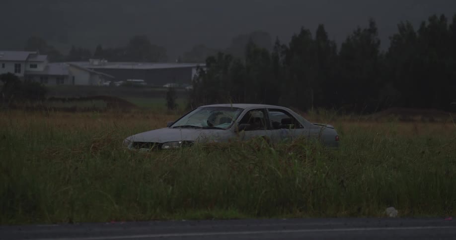 A shot of a dirty gray sedan is seen sitting stationary in a field of tall overgrown grass next to a road as a large white semitruck rapidly passes and momentarily obscures the view