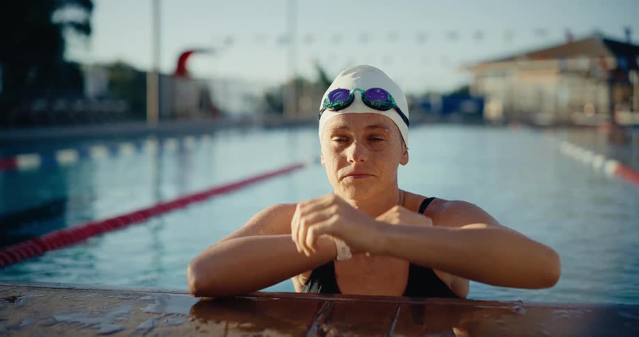 Young adult woman wearing a bathing suit and a swim cap resting on the edge of the pool and removing her googles while panting outdoors during dusk or dawn