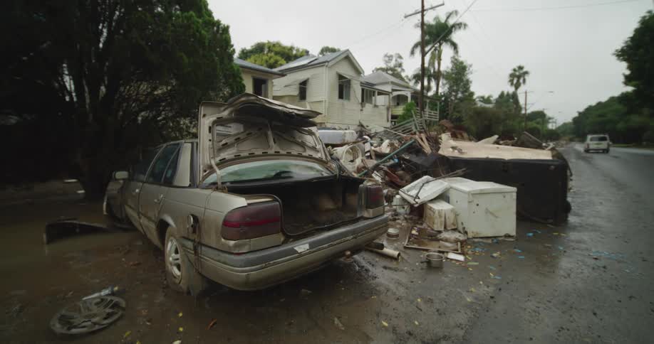 A flood damaged sedan with a crumpled trunk sits on a muddy street next to a large pile of destroyed household items and a residential house in the background