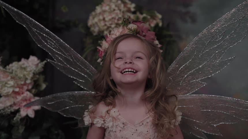 A cheerful young girl dressed in a fairy costume with large translucent wings and a flower wreath laughs happily while facing the camera against a dark floral background