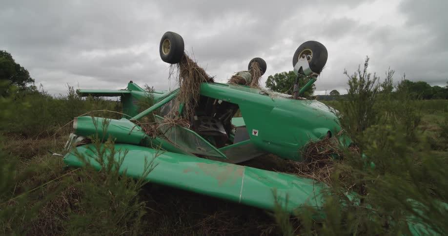 Small green aircraft upside down in grassy area with overcast sky in background