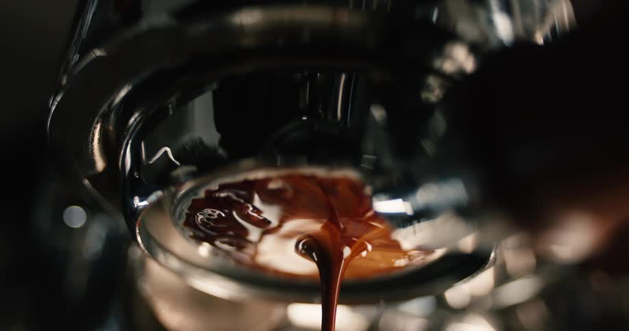 A close view of a coffee machine portafilter shows thick dark espresso slowly dripping and pooling with rich crema before flowing out into a steady stream