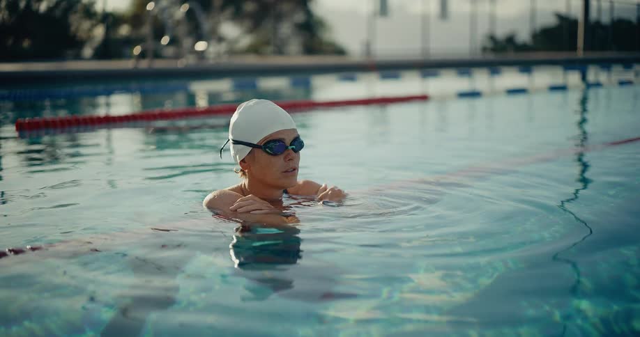 A swimmer wearing a white cap and goggles takes a break at the lane line in an outdoor pool checking a waterproof smartwatch on her wrist during an athletic training session