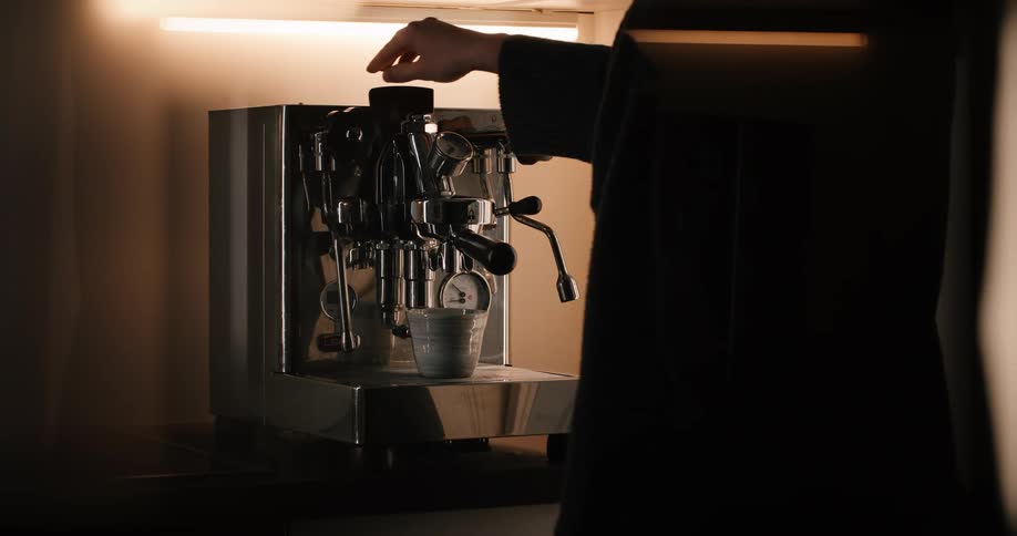 A closeup shot shows a stainless steel espresso machine on a counter illuminated by a strip light above while a person in dark clothing works on the right sidethe person attaches a portafilter to the machine places a small cup underneath and then
