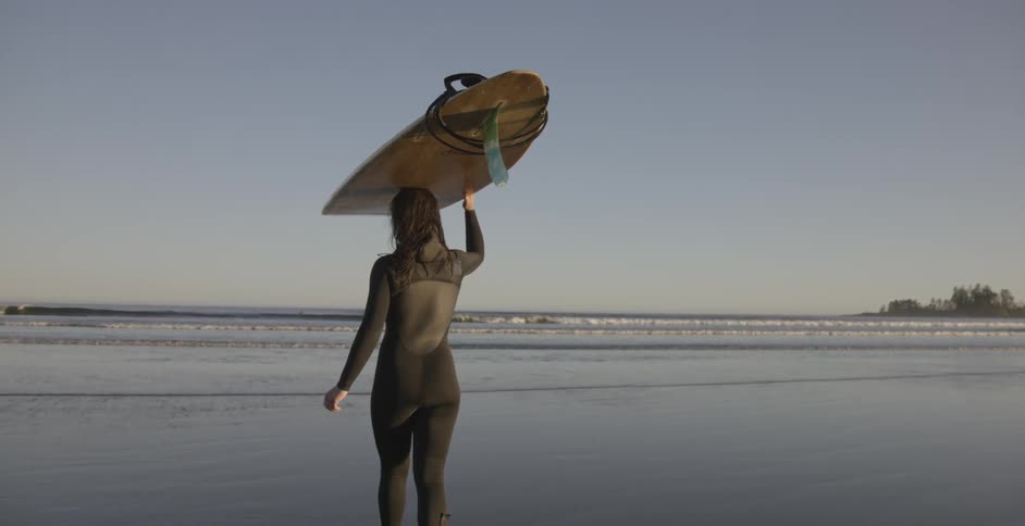 A woman walks on a beach with a surfboard on her head
