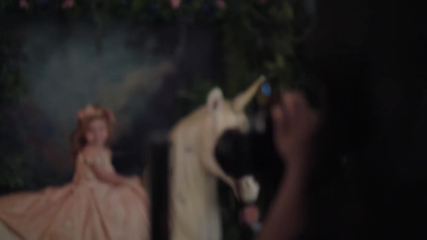 A photographer setting up a scene with a little girl in a pink dress sitting on a white unicorn prop against a dark magical forest backdrop for a fantasythemed photoshoot