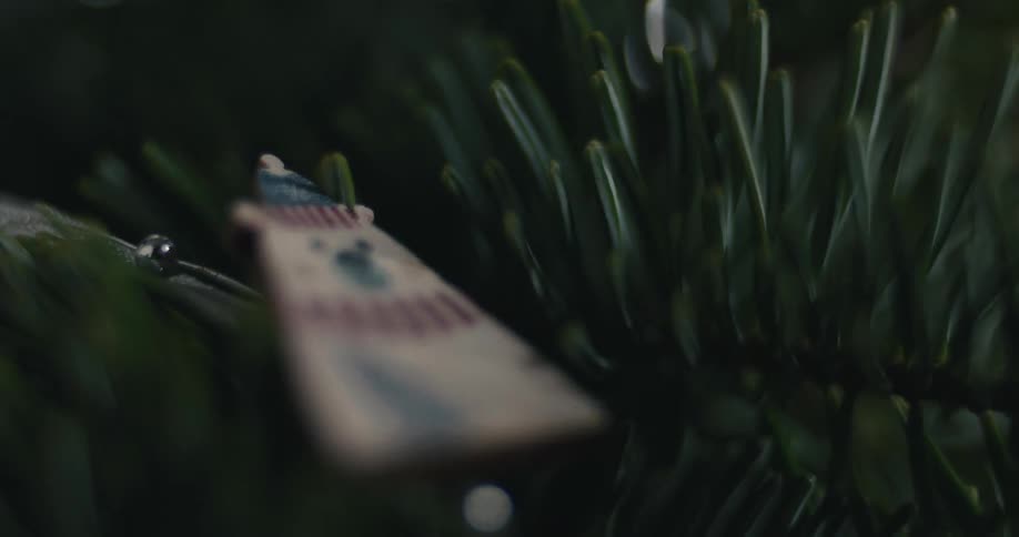 The video is a dark closeup shot focused on a small wooden christmas ornament hanging among the needles of a pine tree or wreath the blurry ornament is shaped like a tag and has a faint pattern of stripes and a stylized christmas tree or snowman