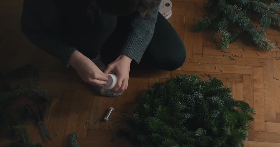 A person sits on the wooden floor next to an evergreen wreath and picks up a white candle to prepare it