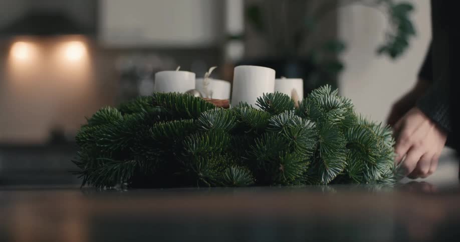 Person placing a festive arrangement with pine branches and white candles on a table indoors