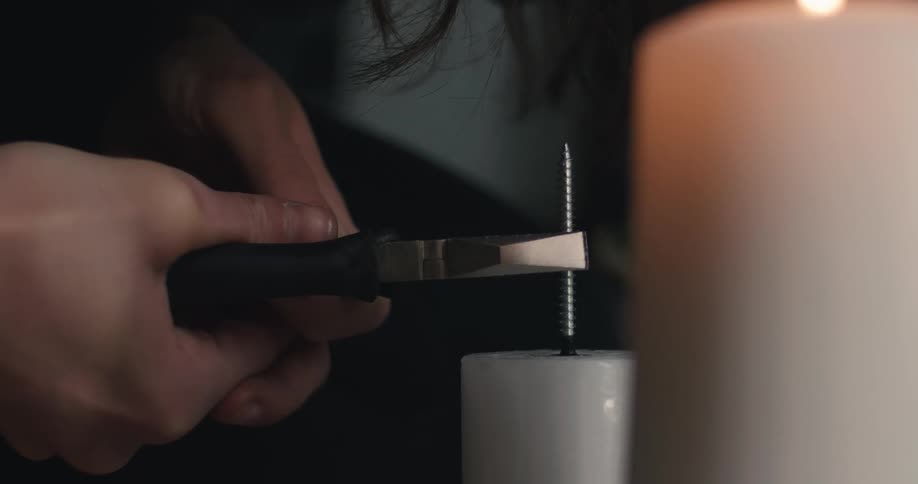 A detailed closeup shows hands holding a tool to press a screw into the top of a white pillar candle with a second lit candle blurred in the background