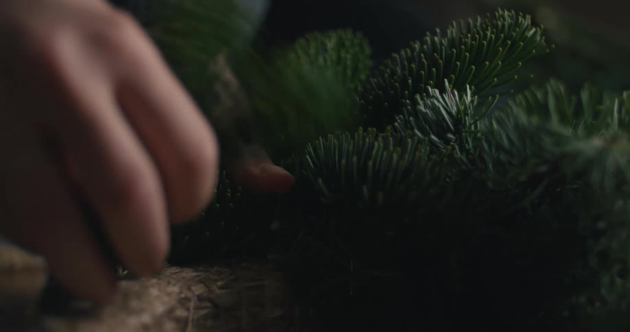 The woman is attaching pine tree limbs to a straw like wreathe